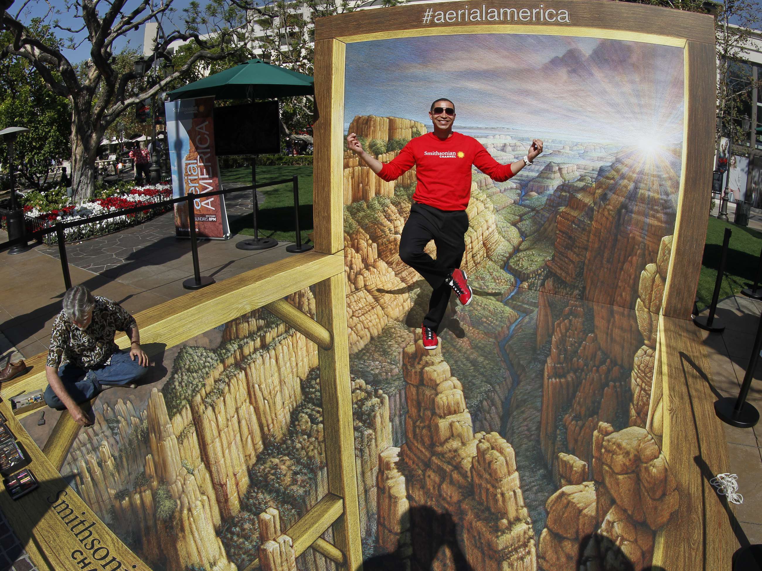 The figure is a sidewalk chalk art of a canyon with various stone pillars. A wooden framework is drawn around three sides of the image. On the fourth side there is an actual wooden framework standing at the end of the image. Within the standing frame is a continuation of the image on the sidewalk with the sky, clouds, and other canyons in the distance painted on it.