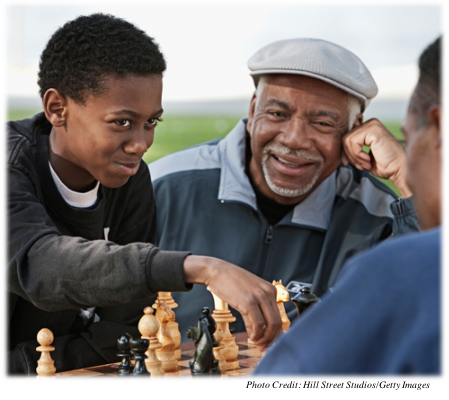 A teenager playing chess with his father.  His grandfather is looking on.